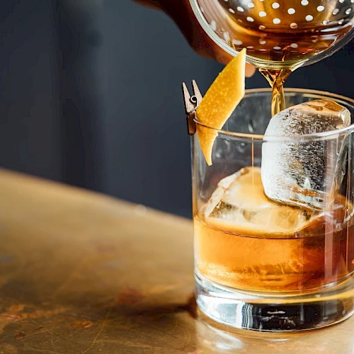 A whiskey tumbler with ice, an orange garnish, and a cocktail being poured from a shaker into a dimly lit bar scene.