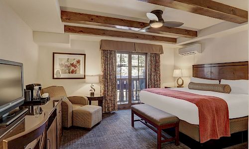A neatly made hotel room with a large bed, chairs, window with curtains, and a ceiling fan.