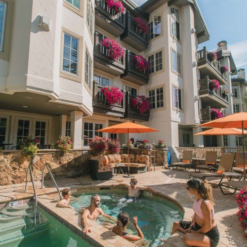 Resort pool with several people lounging and kids swimming by a multi-story building adorned with pink flowers on balconies and orange umbrellas.