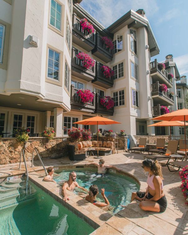 Resort pool with several people lounging and kids swimming by a multi-story building adorned with pink flowers on balconies and orange umbrellas.