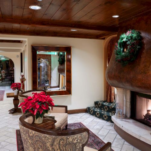 A cozy living room with a stone fireplace, wooden ceiling beams, festive poinsettias, a mirror and art on the walls, and comfortable seating.