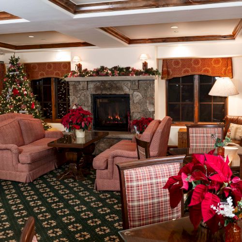 A cozy living room with a stone fireplace, festive Christmas tree, and red poinsettia centerpieces; pink and plaid seating, warm lighting, and green patterned carpet.