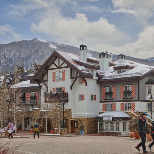 A quaint alpine village with chalet-style houses, snow-capped mountains, and a sunny, clear sky.