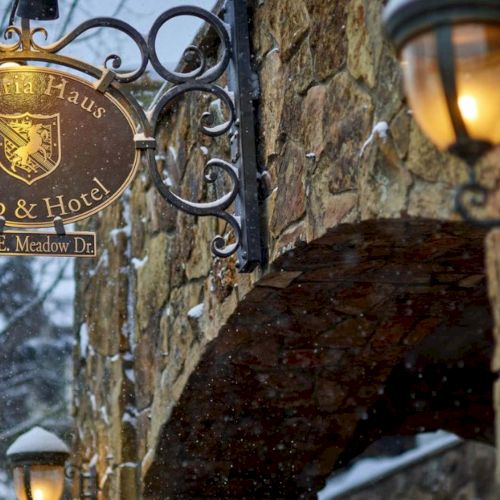 A rustic stone hotel sign reading &ldquo;R&ouml;sler House Club & Hotel&rdquo; hanging by an ornate wrought-iron bracket near a warmly lit stone arch in a snowy scene.