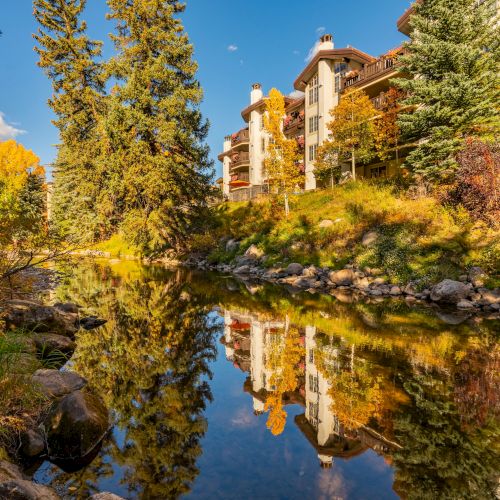 Autumn apartment buildings rise beside a pond, with gold trees and crisp reflections under a bright blue sky.