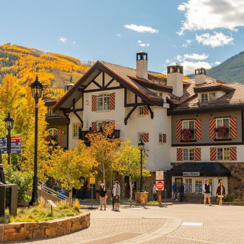 Alpine lodge with red-and-white shutters, autumn foliage, mountains, and pedestrians in a sunny resort courtyard.