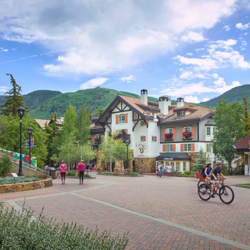 A sunny mountain village plaza with a chalet-style building, trees, lampposts, pedestrians and cyclists, framed by green hills.