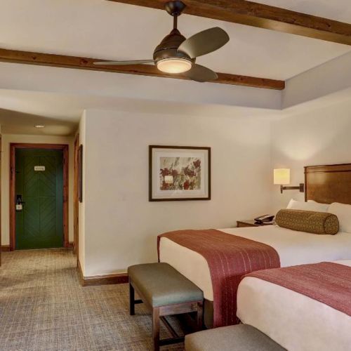 A hotel room with two queen beds, wooden headboards, a ceiling fan, desk, TV, lamps, a bench, and an entrance to a hallway&mdash;bright and tidy.