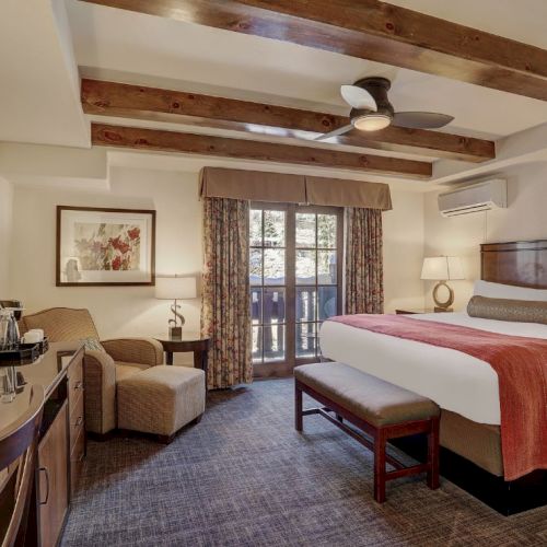 A hotel room with a king bed, bench, seating area, desk, and balcony doors under exposed wooden beams, cozy and well-lit.