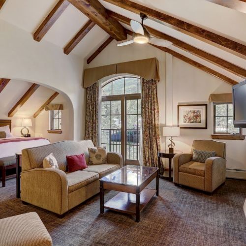 A cozy hotel suite with a living area, exposed wooden beams, a fireplace, a large window, and a bed visible in the background, all in warm tones.