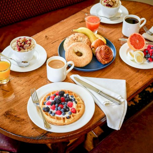 Brunch spread on a wooden table: fruit-topped waffle, pastries, juice, and coffee with berries and citrus.