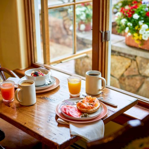 A cozy breakfast by a sunlit window: coffee, orange juice, assorted pastries on a wooden table, with flowers outside and warm decor.