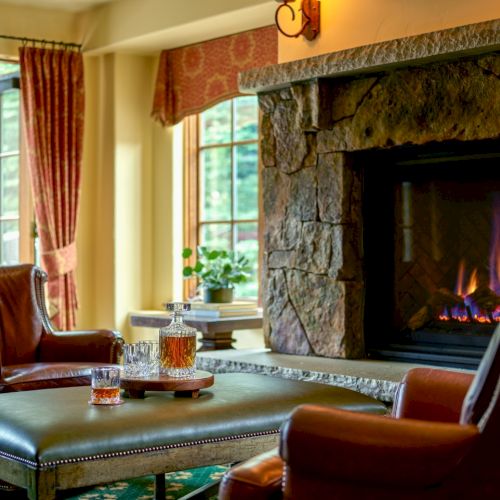A cozy living room with a stone fireplace, leather chairs, a coffee table, natural light from open windows, and warm ambient glow.