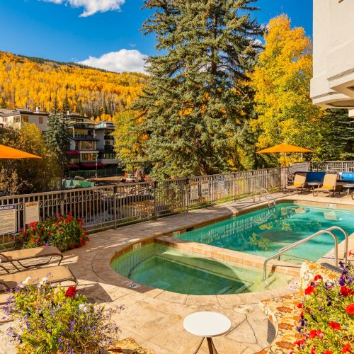A sunny mountain resort pool with orange umbrellas, lounge chairs, and autumn trees in golden leaves behind it.