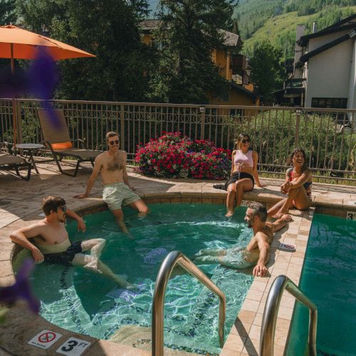 A backyard pool scene with lounge chairs, a shade umbrella, and people enjoying a sunny day by a fence and trees.