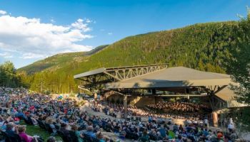 An outdoor amphitheater filled with a large audience watching a live concert against a backdrop of green hills under a bright blue sky.