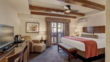 A hotel room with two queen beds, a bench at the foot, a seating area, a desk, flat-screen TV, ceiling fans, exposed wooden beams, and a balcony door with curtains.