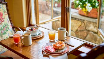 Breakfast on a wooden table by large windows&mdash;fruit, toast/cake, juice, and coffee with a colorful garden view outside.