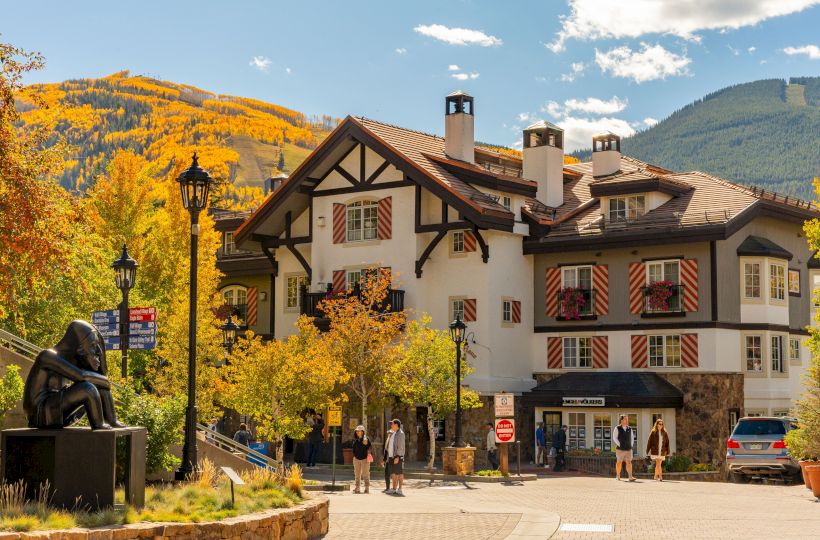 A Swiss-style inn with black-timber beams and flags, autumn trees, people, and mountains under a blue sky.