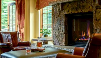 A cozy living room with a stone fireplace lit by a fire, leather chairs, a table with a bottle and glasses, and red curtains, by a window.