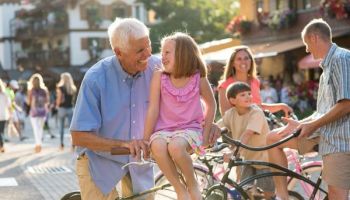 An elderly man smiles at a girl on a bicycle while a crowd chats and rides by in a sunny outdoor square.