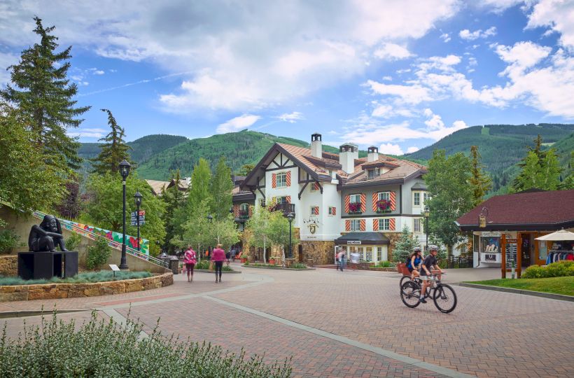 A charming alpine village square with a white cottage-style inn, cyclists, pedestrians, lush greenery, and mountains under a blue, partly cloudy sky.