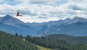 A person in a red outfit ziplining high above a forested mountain valley, with a bright blue sky and scattered clouds.