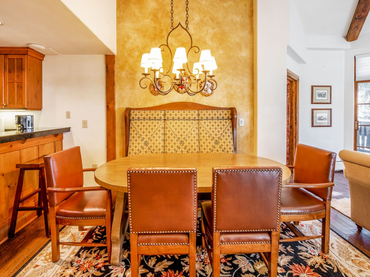 Warm dining room with a large chandelier, round wooden table, leather chairs, patterned banquette, and a glimpse of kitchen cabinetry.