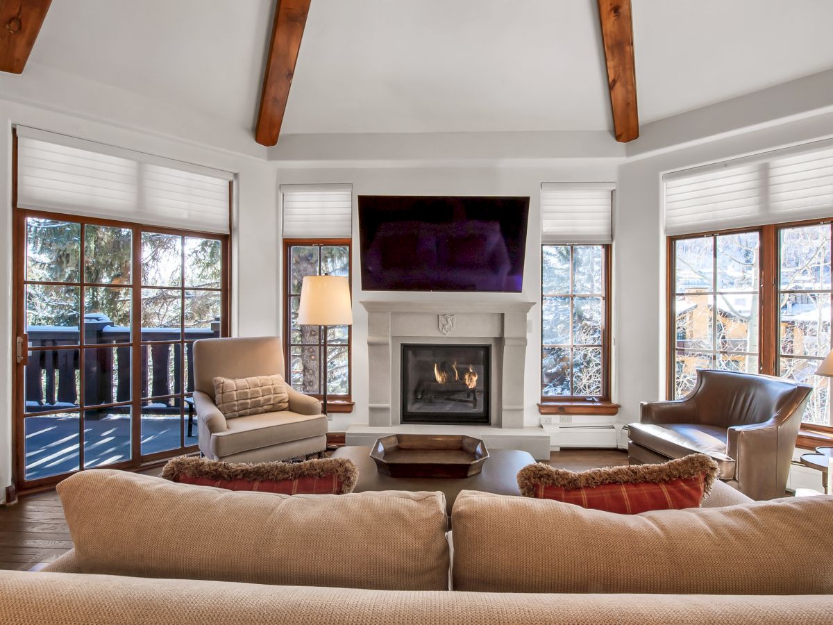 A cozy living room with a fireplace, wall-mounted TV, large windows, and beige seating under wood ceiling beams and blinds,