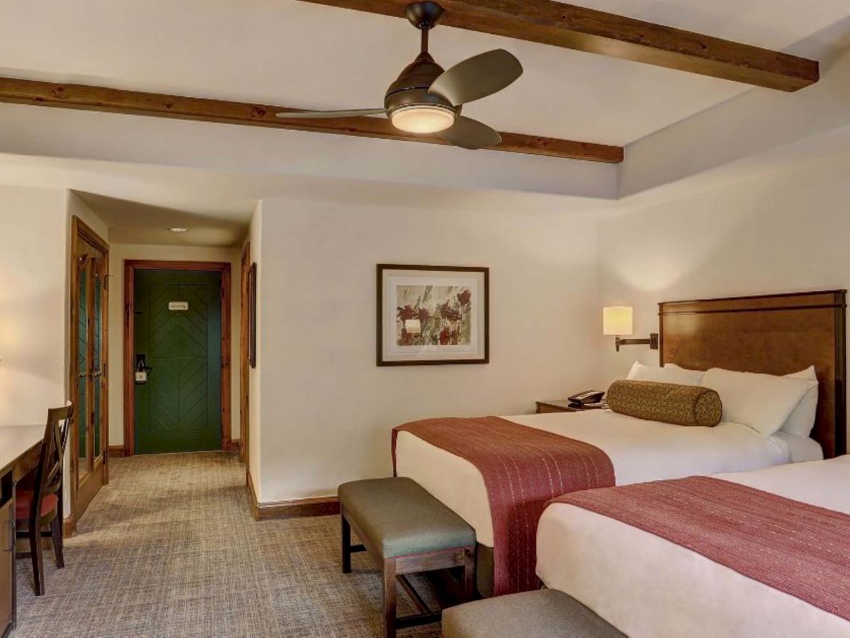 A hotel room with two double beds, wooden headboards, a bench, desk with chair, a TV, carpet, ceiling fans and an entry hallway to a green door.