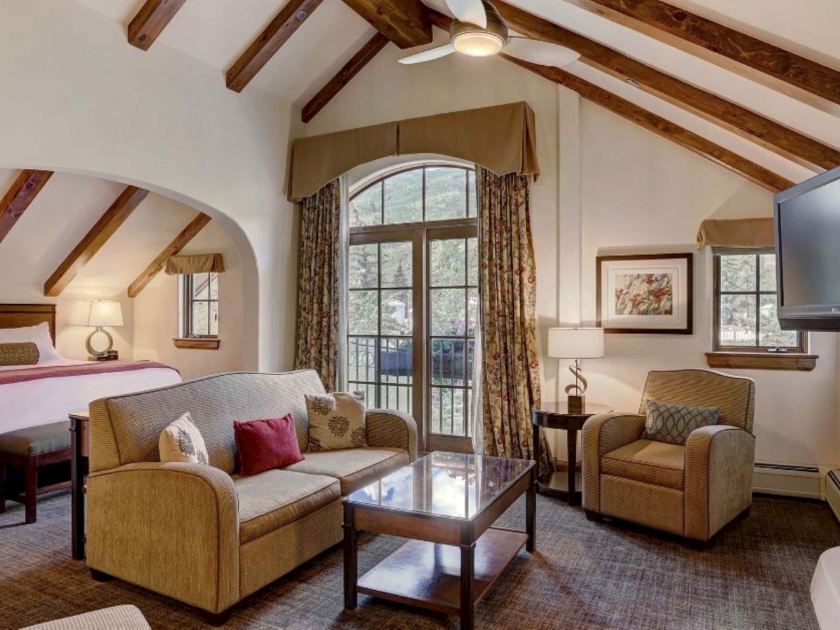 A cozy hotel suite with a living area,arbored ceiling beams, a fireplace, flat screen, sofa, armchairs, coffee table, and a balcony door.