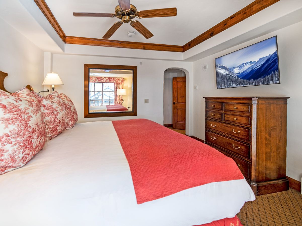 A cozy bedroom with a ceiling fan, red-and-white bedspread, dresser, framed window, and mountain landscape wall art.