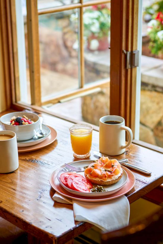 A cozy breakfast table by a sunny window with coffee, juice, toast, pastries, and fresh flowers outside, inviting a peaceful morning.
