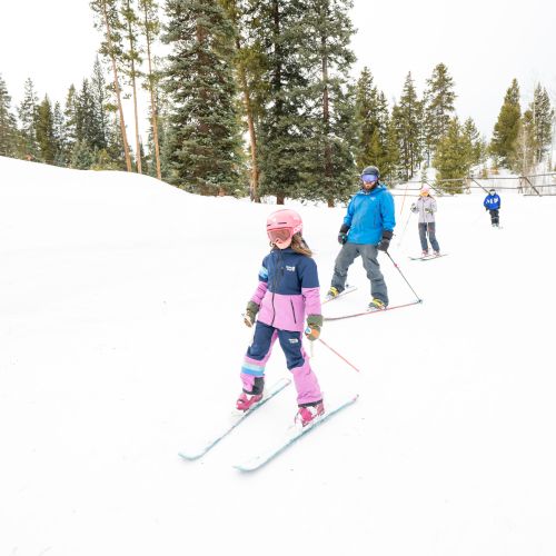 Two kids ski down a snowy slope with an instructor in the background, wearing colorful winter outfits among a frosty forest.
