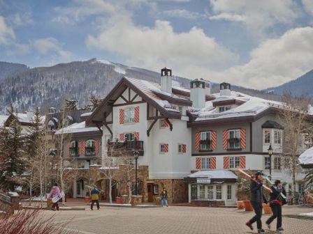 A snowy mountain village with a chalet-style hotel, pine trees, and people strolling along a paved square under a partly cloudy sky.