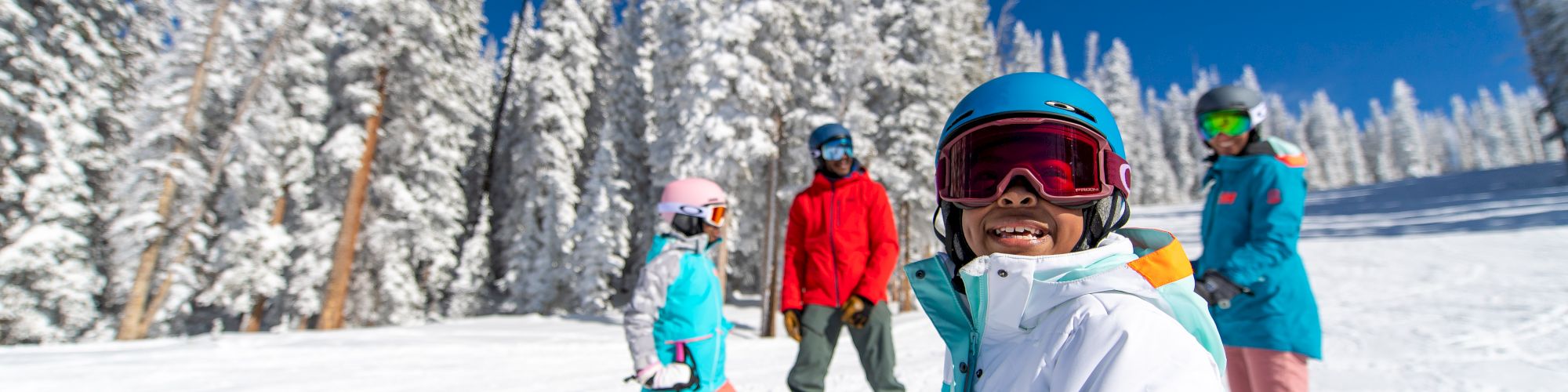 A group of kids skiing on a sunny, snowy slope with snow-covered trees in the background; blue skies, wearing colorful jackets and helmets.
