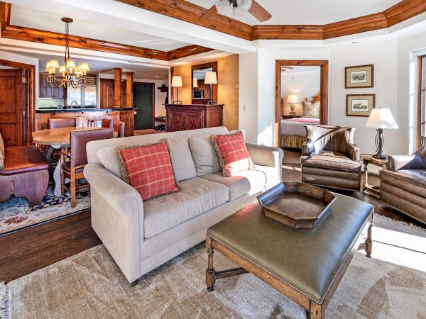 A spacious, elegantly furnished living room with a beige sofa, brown armchairs, a large ottoman, wooden ceiling beams, and a dining area in the background.