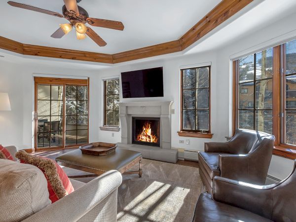 A bright living room with a stone fireplace, ceiling fan, large windows, a TV above the mantel, and comfy seating around a coffee table.
