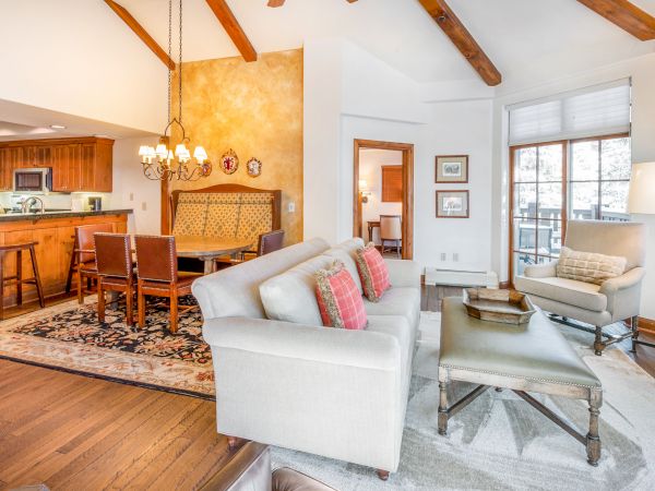 Cozy open-concept living area with a white sofa, wooden ceiling beams, a kitchen bar, dining area, and a sunny seating nook by large windows.