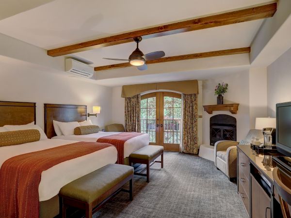 The image shows a hotel room with two beds, a ceiling fan, TV, fireplace, and a balcony door with natural light streaming in.
