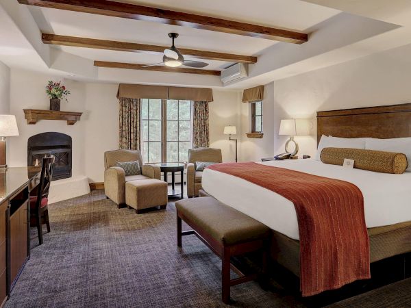 A spacious hotel room with a large bed, TV, seating area, desk, and fireplace. Neutral tones and natural light create a cozy ambiance.