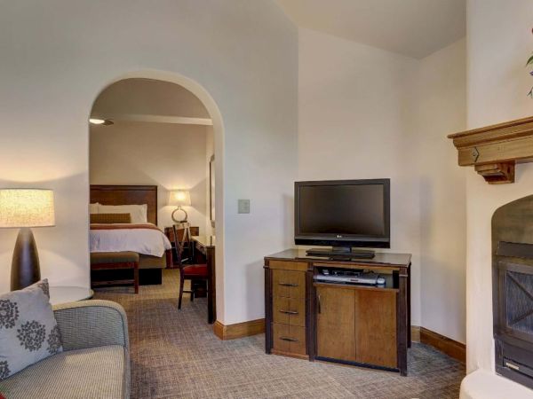 A cozy living area with a fireplace, TV, couch, and pillows, leading to a bedroom with a bed, lamp, and wooden furniture.