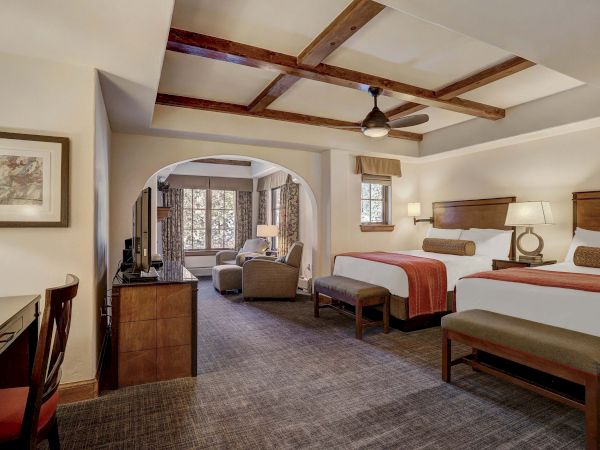 The image shows a spacious hotel room with two beds, a TV, desk, and seating area under a beamed ceiling and warmly lit lamps.