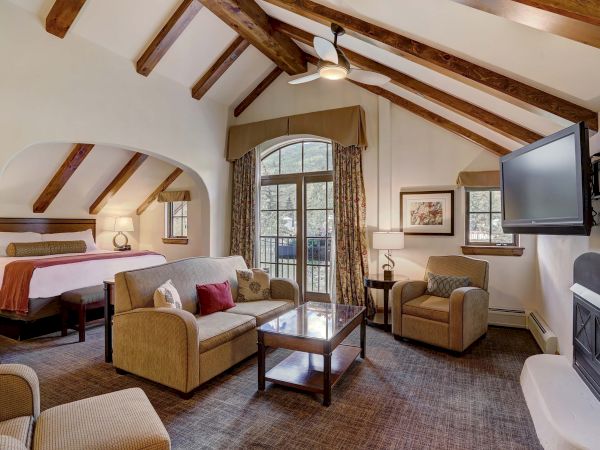 A cozy living space features a bed, sofa, armchairs, a fireplace, and a mounted TV with a wooden-beam ceiling and large windows.