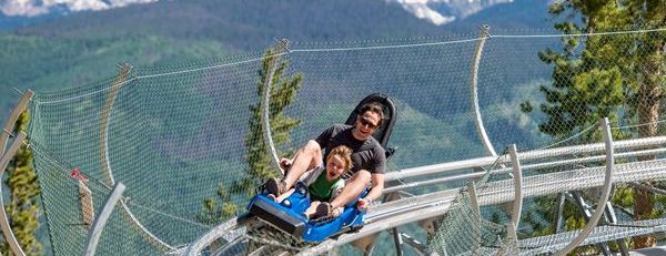 Two people ride a rollercoaster-like track along a mountainside, enjoying the scenic view as they zoom by.