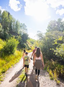 A family hike on a sunny dirt trail through a green, wooded hillside, with bright skies and long shadows as they walk away.