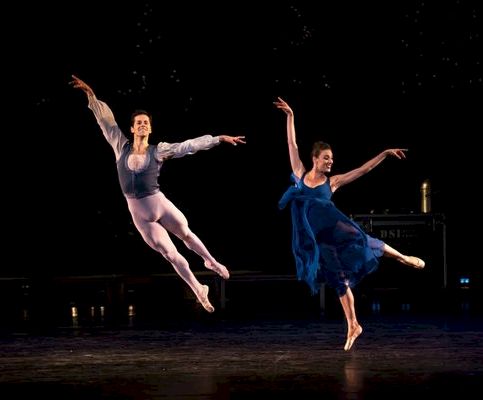 Two dancers leap and stretch on a dark stage, illuminated by a single spotlight, frozen in a graceful mid-air pose.