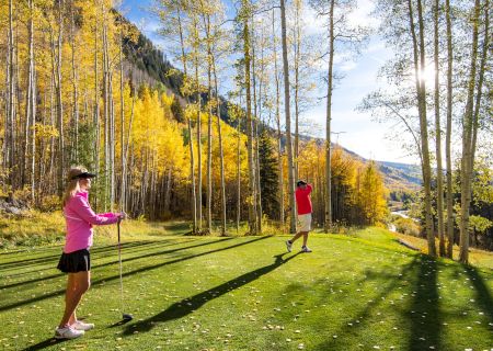 Two hikers-golfers stroll a sunny autumn fairway lined with tall aspens, yellow leaves fluttering as they play.