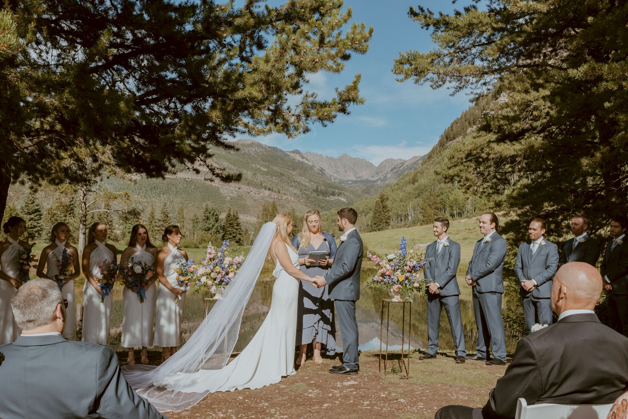 A bride and groom exchange vows outdoors, surrounded by bridal party in silver dresses and gray suits, with mountains and trees in the background.