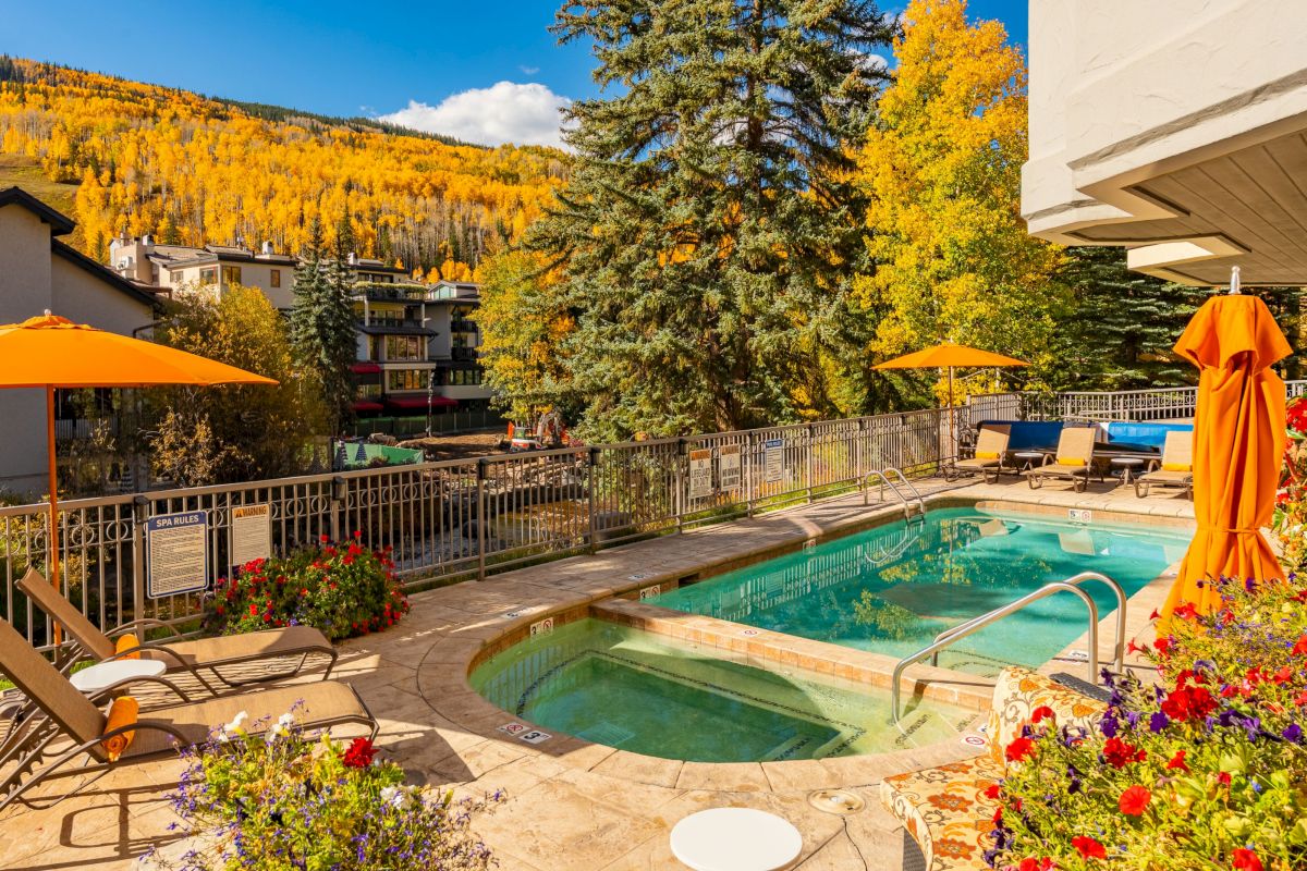 Outdoor resort pool with sun loungers and orange umbrellas, backed by autumn-gold trees and mountains under a blue sky.